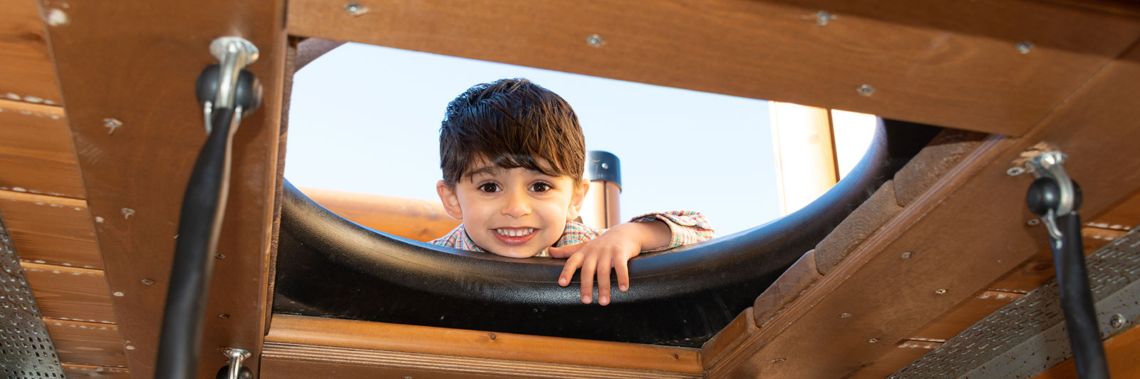 A young child looks through a gap from within a wooden playground unit, he is looking directly at the camera and widely smiling.